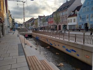 Foto vom offengelegten Fluss in der Stadt Freising mit Sitzbänken und Steinen im Wasser, darum Häuser mit Geschäften.
