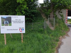 Schild mit Ankündigung des Neubaus einer Unterführung unter einer Bundesstraße in Oberbayern. Rechts am Bildrand ist zu sehen, dass in der aktuellen Unterführung einer Landstraße unter der BAB A8 ein minimaler Streifen für Radfahrer und Fußgänger frei ist.